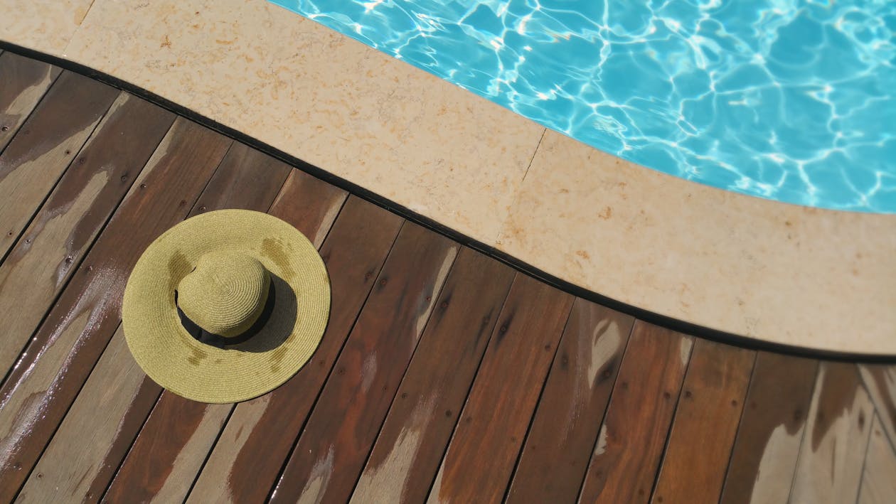 A straw hat rests on a wooden deck beside a sparkling blue swimming pool, perfect for summer relaxation. — pool deck coating and resurfacing